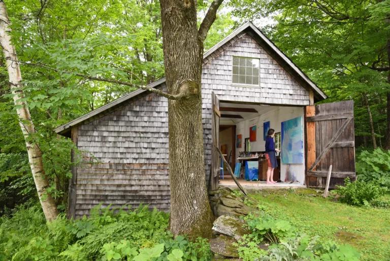 Photograph of a brown shingled barn nestled in lush, green woods, with the double-doors open; inside reveals a light-skinned woman working on a blue abstract painting.