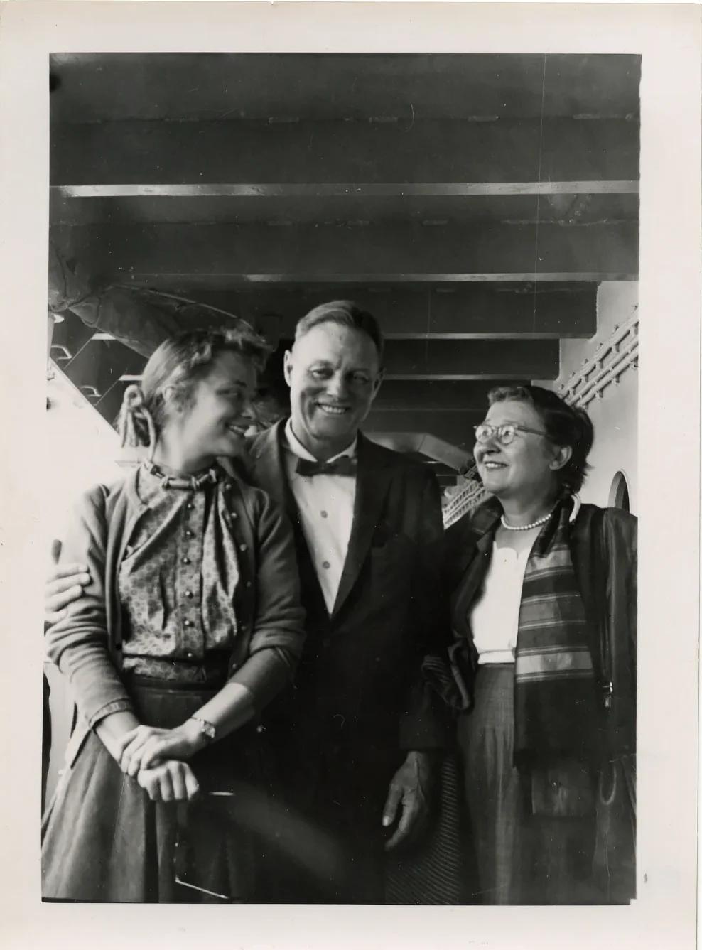 Black and white photograph of a man, young woman, and an older woman on a ship, all with light skin tones and smiling. The two women are looking at each other.