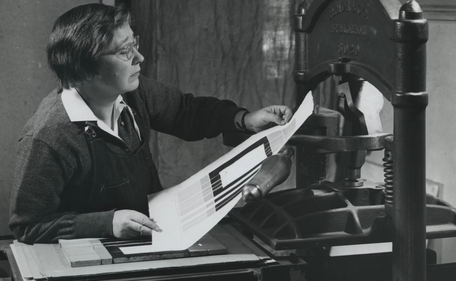 Black and white photograph from 1954 of women a woman with pale skin, examining a print just pulled off of a large printmaking press.