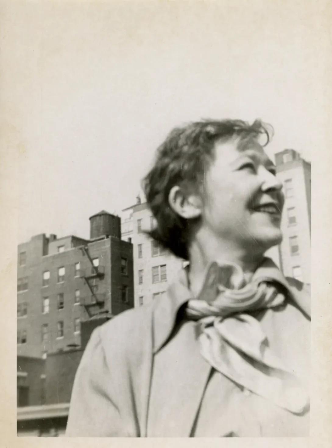 Black and white portrait of a woman with a light skin tone, looking off to the right and smiling; NYC apartments are seen behind her.