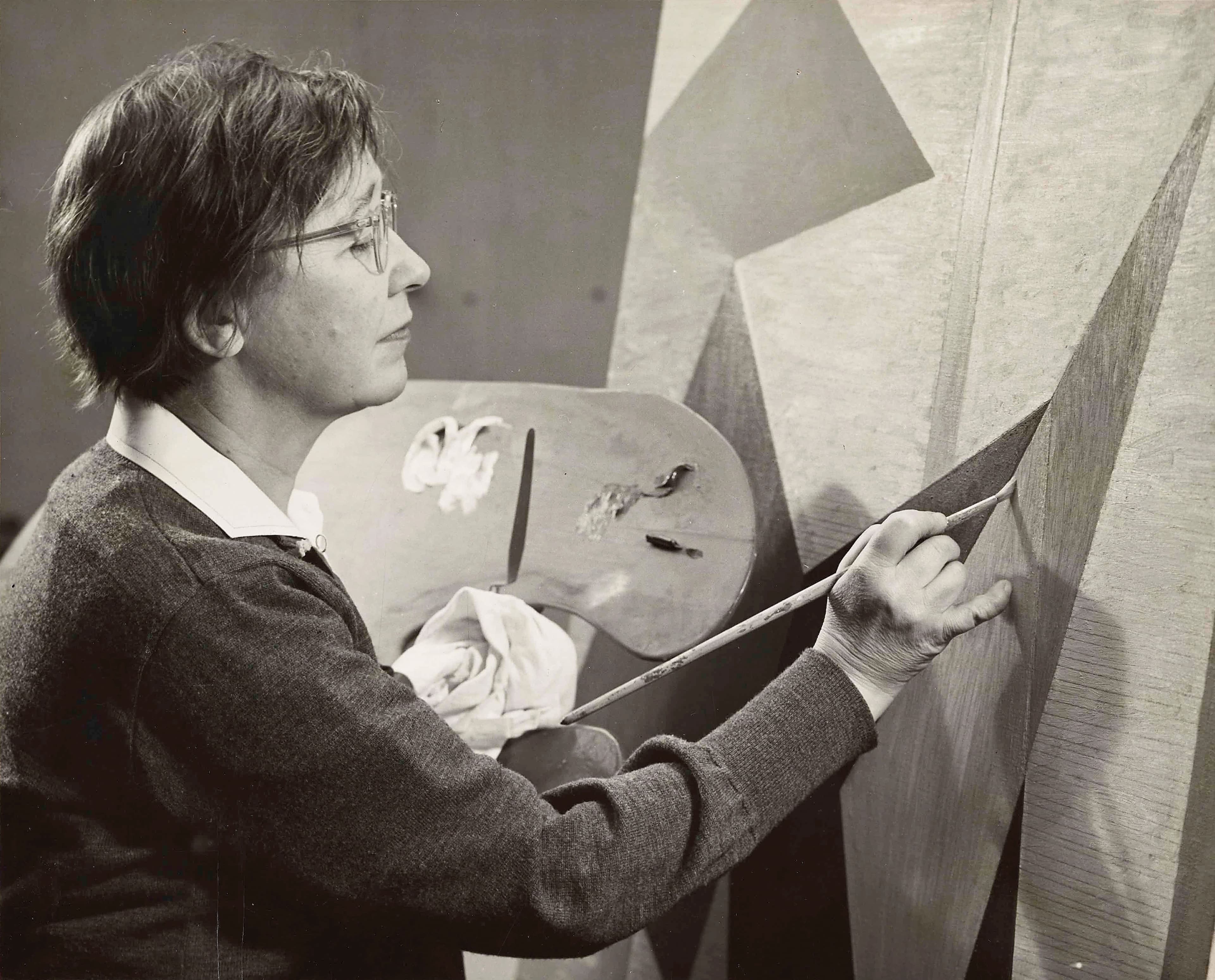 A black and white photograph of a light-skinned woman, holding a palette in her left hand and a paint brush on the right; she is in the process of creating an abstract painting.