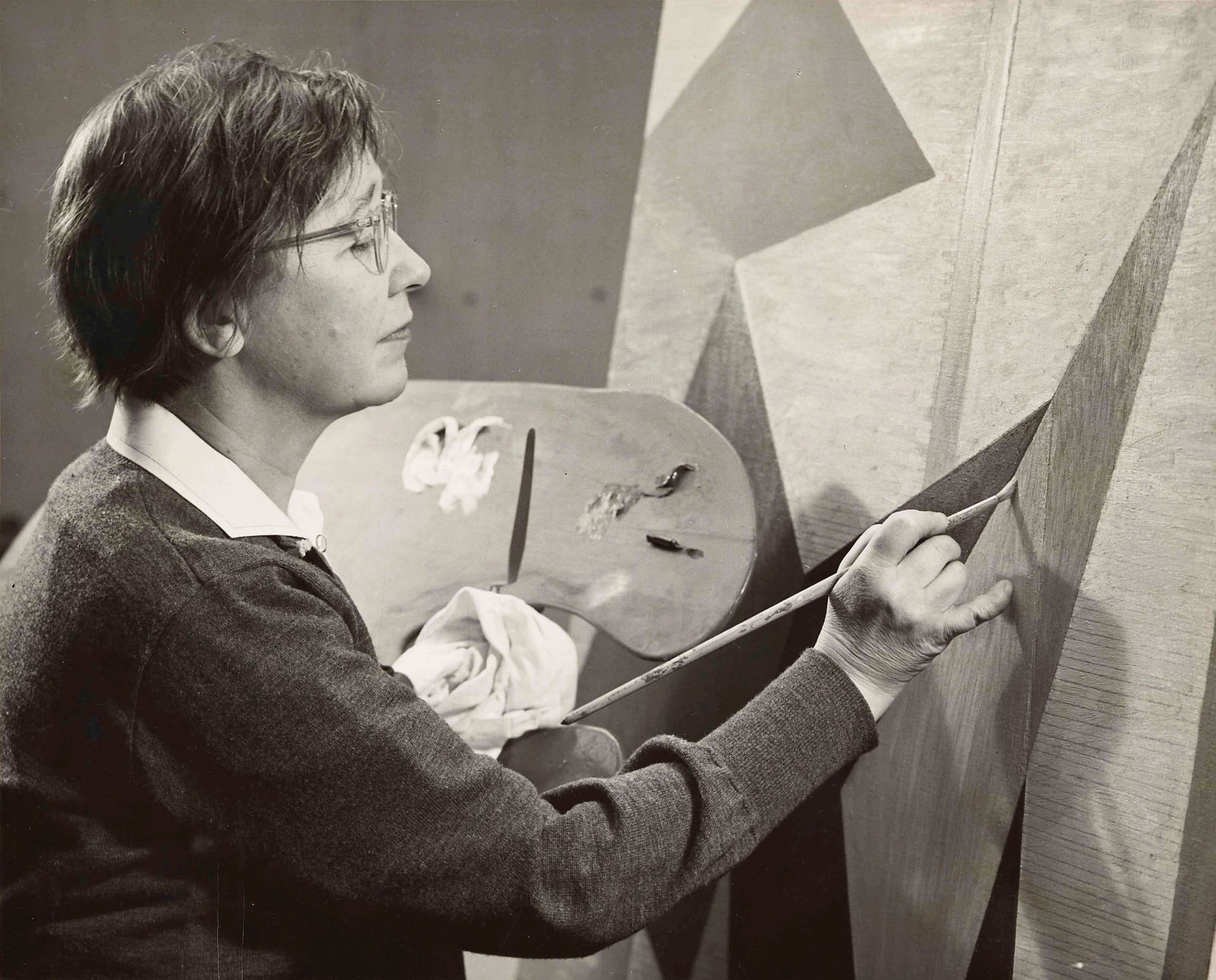 A black and white photograph of a light-skinned woman, holding a palette in her left hand and a paint brush on the right; she is in the process of creating an abstract painting.