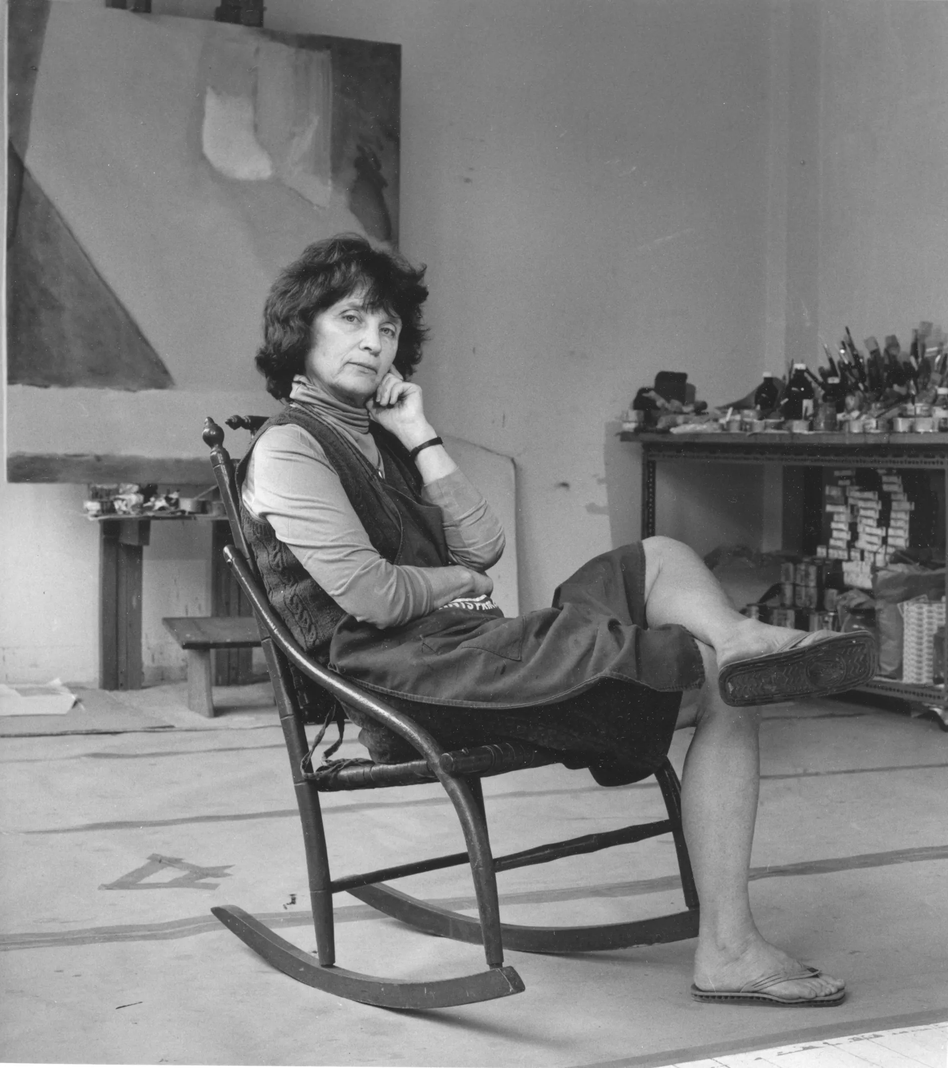 Emily Mason in her NYC studio, phot by Tommy Naess, 1991 Black and white photograph of a light-skinned woman with short hair, sitting a rocking chair in a painting studio.