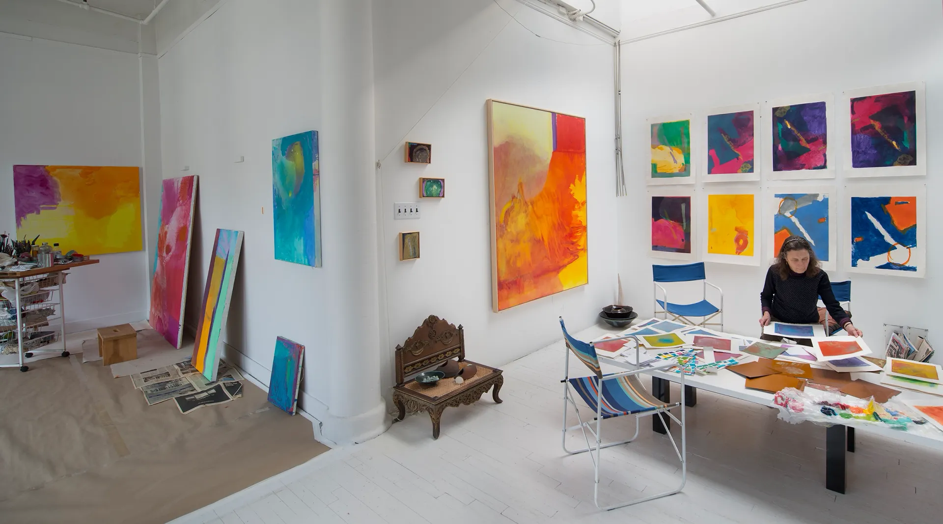 Emily Mason, 20th Street, 2015 A photograph of an all-white, large room with high ceilings; the walls have colorful, abstract paintings and prints hung on the walls, and an older woman with a light skin tone looks through prints spread out on a table.